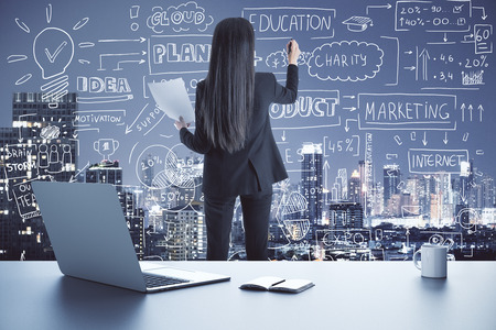 Back view of young businesswoman drawing creative business sketch on abstract city background with charts on laptop screen and coffee cup. Report and finance conceptの写真素材