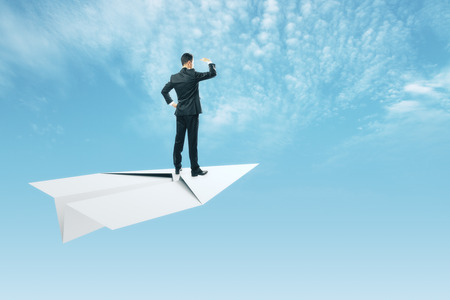 Young businessman looking into the distance while standing on white paper plane. Sky and clouds background. Freedom and research conceptの写真素材