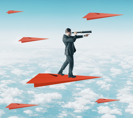 Young businessman using telescope to look into the distance while standing on red paper plane on sky background. Vision and direction conceptの写真素材