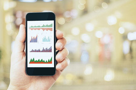 Businessman hand holding smartphone with business charts on blurry bokeh interior background. Technology and finance conceptの写真素材