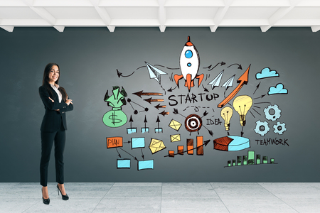 Attractive young european businesswoman standing in concrete office interior with start up sketch on wall. Startup and investment conceptの写真素材