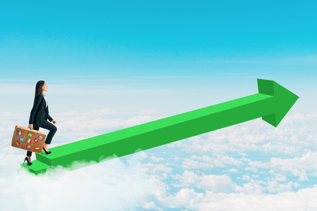 Side view of young businesswoman with travel suitcase walking on green arrow on sky background. Success and career conceptの写真素材