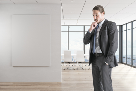 Thoughtful young european businessman standing in modern office interior with empty poster. Boss and research concept.の写真素材