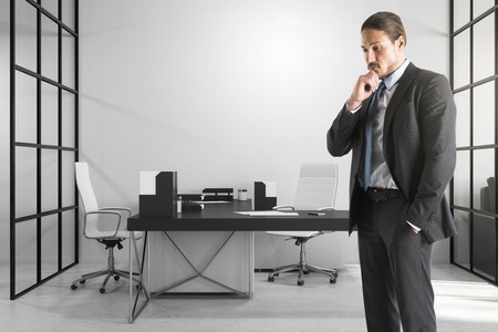 Thoughtful young european businessman standing in modern office interior. Boss and research concept.の写真素材