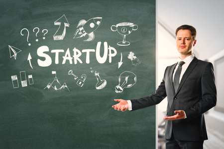 Attractive young businessman standing in office interior with startup sketch on chalkboard. Start up and presenation conceptの写真素材