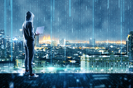 Young hacker with laptop standing on abstract glowing city rooftop background with binary code background. Hacking and phishing concept. Double exposureの写真素材