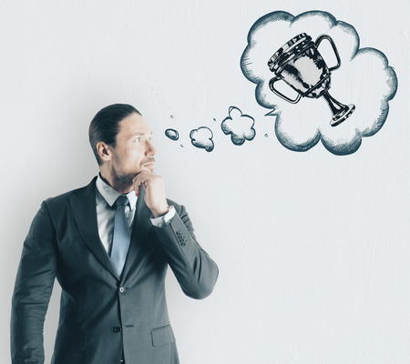 Attractive young businessman thinking about trophy on concrete wall background. Leadership and success conceptの写真素材