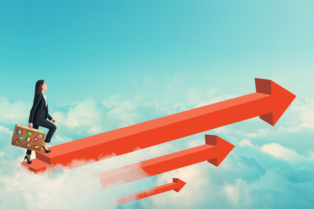 Side view of young businesswoman with travel suitcase walking on red arrows on sky background. Success and career conceptの写真素材