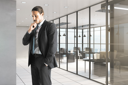 Portrait of attractive thoughful young european businessman standing in contemporary office interior. Research and executive concept.の写真素材