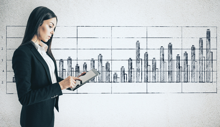 Side view of attractive young businesswoman using tablet on concrete wall background with business chart sketch. Finance and mobile conceptの写真素材