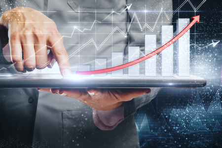 Close up of hand using tablet with business chart hologram on blurry office interior background. Trade and finance concept. Double exposureの写真素材