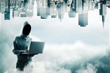 Hacker in hoodie using laptop on abstract sky background with clouds and upside-down city skyline. Technology and hacking conceptの写真素材