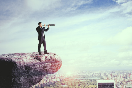 Side view of young businessman on cliff using telescope to look into the distance on city background. Research and research conceptの写真素材