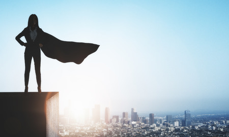Backlit superhero businesswoman with waving cape standing on rooftop on city and sky background. Copy space. Leadership and success conceptの写真素材