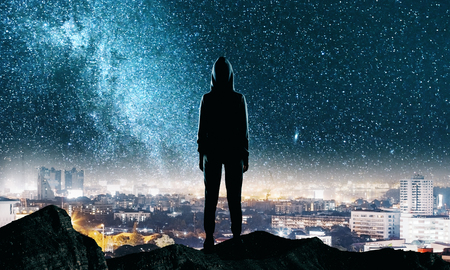Back view of hacker standing on top of hill with night starry sky and city skyline background. Future and success conceptの写真素材