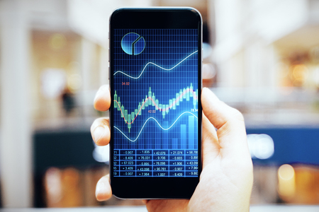 Close up of hand holding smartphone with forex chart on blurry background. Trade and mobile gadget conceptの写真素材