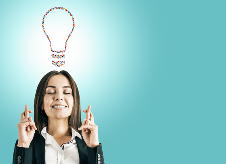 Portrait of attractive young european businesswoman with crossed fingers and creative hand gesture lightbulb on blue background. Communication, community, inspire, idea and innovation conceptの写真素材