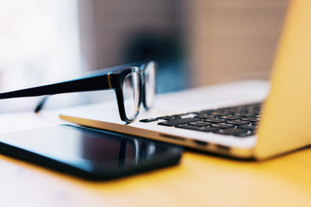 Close up of eyeglasses, smartphone and laptop keyboard on wooden desktop. Blurry background. Workplace and software conceptの写真素材