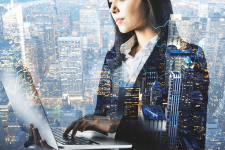 Side view of attractive young businesswoman using laptop on blurry night city background. Technology and network concept. Double exposureの写真素材