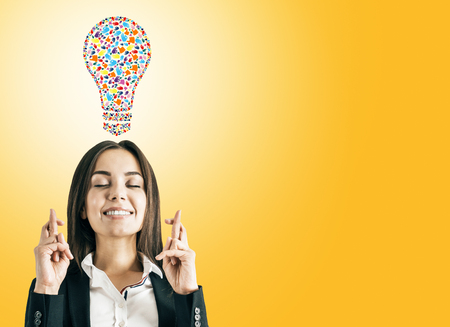 Portrait of attractive young european businesswoman with crossed fingers and creative hand gesture lightbulb on orange background. Communication, community, inspire, idea and innovation conceptの写真素材