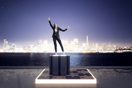 Happy young businesswoman on puzzle pedestal. Blurry city skyline background. Quiz and solution conceptの写真素材