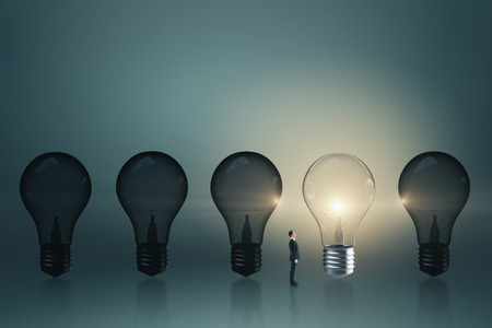 Tiny businessman looking at row of light bulbs with one illuminated. Success and leadership concept.の写真素材