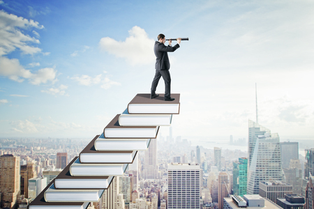 Side view of young businessman with telescope on creative book ladder. New York city skyline background. Growth, education and future conceptの写真素材