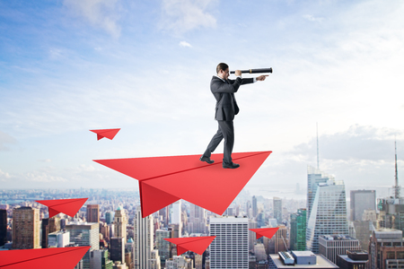 Young businessman with telescope standing on abstract red paper plane on sky and city background. Future and leadership conceptの写真素材