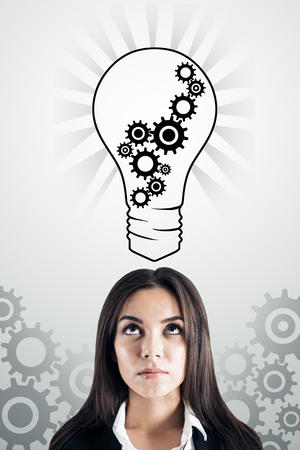 Pretty thoughtful young businesswoman with creative cogwheel light bulb sketch on subtle white background. Idea, innovation and machinery conceptの写真素材