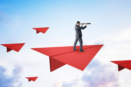Young businessman on red paper plane looking into the distance using binoculars. Vision and research conceptの写真素材