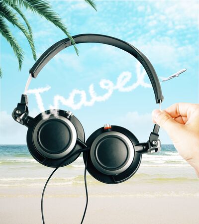 Hand holding headphones on creative beach background with airplane and text in sky. Music and travel conceptの写真素材