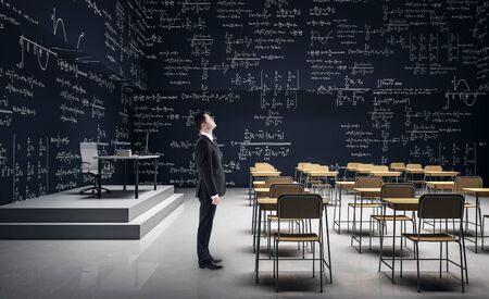 Thoughtful man standing in modern classroom interior with furniture and mathematical formulas on wall. Math and complex algorithm concept.の写真素材