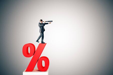 Side view of young businessman standing on red percent sign and looking into the distance on grey background. Interest rate, investment and loan conceptの写真素材