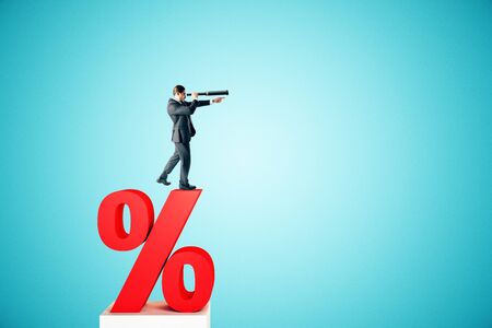 Side view of young businessman standing on red percent sign and looking into the distance on blue background. Interest rate, investment and loan conceptの写真素材