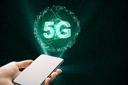 Close up of hand holding smartphone with creative glowing 5G internet hologramn on dark background. Technology and speed conceptの写真素材