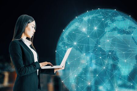 Attractive youong caucasian businesswoman using laptop with glowing polygonal globe on blurry night city background. AI and data concept. Multiexposureの写真素材