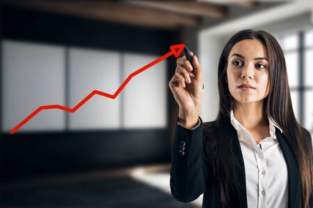 Attractive young european businesswoman drawing upward red economic arrow on blurry office interior background. Growth and increase concept. Multiexposureの写真素材