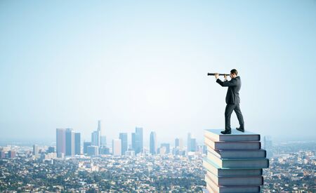 Side view of young businessman standing on books and using telescope to look into the distance on blue sky and city background. Research, education and vision conceptの写真素材