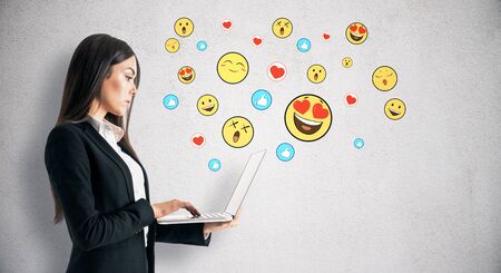 Portrait of attractive young businesswoman using laptop with emotive smileys on subtle concrete background. Communication and emotion conceptの写真素材