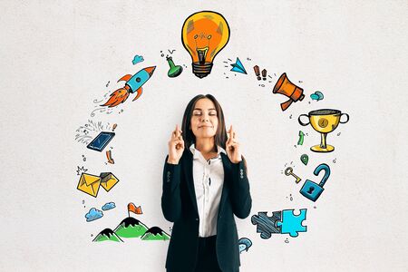 Attractive young european businesswoman with crossed fingers standing on concrete wall background with round business sketch lamp. Idea and success conceptの写真素材