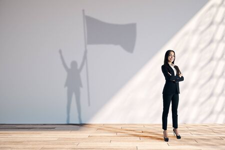 Happy young european businesswoman with flag shadow in white interior with sunlight. Leadership and success concept.の写真素材