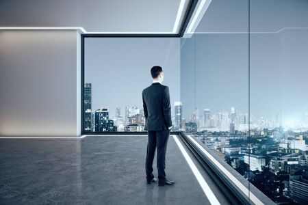 Businessman looking out of window in contemporary empty spacious office interior with illuminated panoramic city view. Research concept.の写真素材