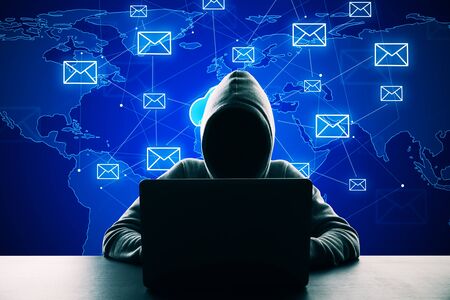 Hacker in hoodie using laptop at desktop with creative glowing cloud email map network hologram on blue background. Cloud computing, newsletter and interactive hud conceptの写真素材