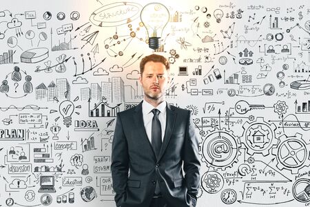 Thoughtful young businessman with glowing light bulb and business sketch on concrete wall...