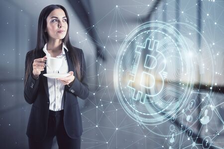 Finance and cryptocurrency concept. Portrait of attractive young european businesswoman drinking coffee with glowing bitcoin hologram on blurry office interior. Multiexposureの写真素材