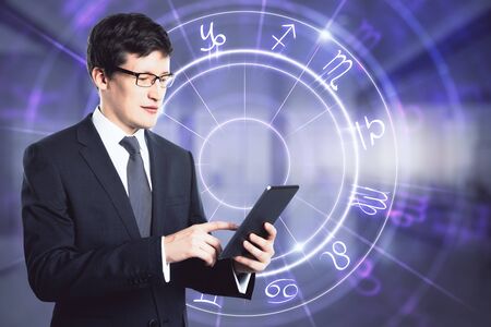 Forecast and fortune telling concept. Portrait of attractive young european businessman with tablet and glowing globe astrological calendar hologram interface on blurry office interior. Multiexposureの写真素材