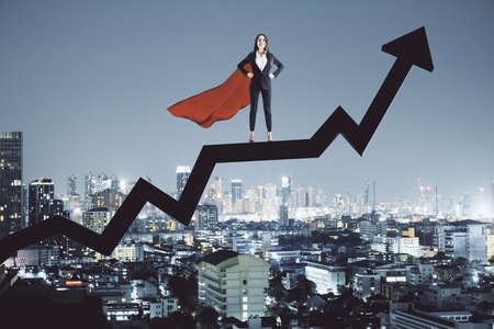 Businesswoman in hero's cloak standing on black arrow on city background. Success and startup concept.の写真素材