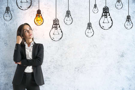 Businesswoman thinking and drawing yellow lamps on gray concrete wall. Innovation and leadership conceptの写真素材