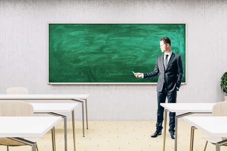 Businessman standing in modern classroom with empty chalkboard, furniture and daylight. Education and school concept. Mock up,の写真素材