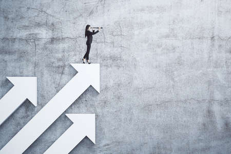 Young businesswoman standing on white arrow and using telescope on concrete wall background. Business and challenge concept.の写真素材